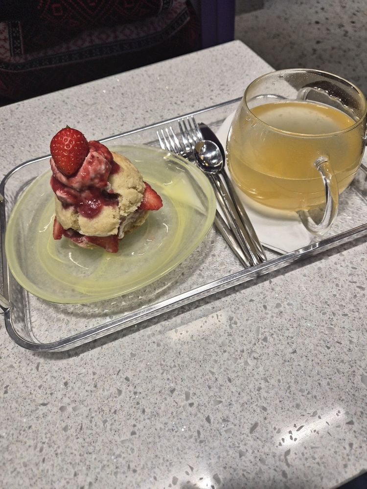 A strawberry cream cake slice on a plate, served on a metal tray beside a glass cup of tea.