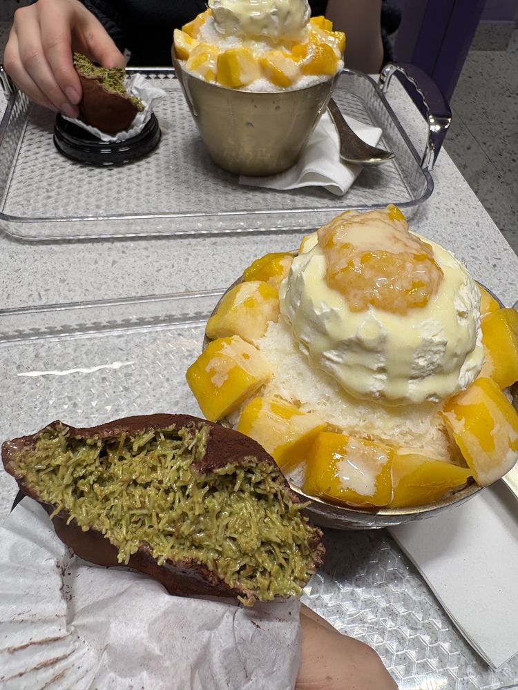 A table of desserts including a mango bingsu, a matcha cake slice and a gold cup, with a hand reaching in.