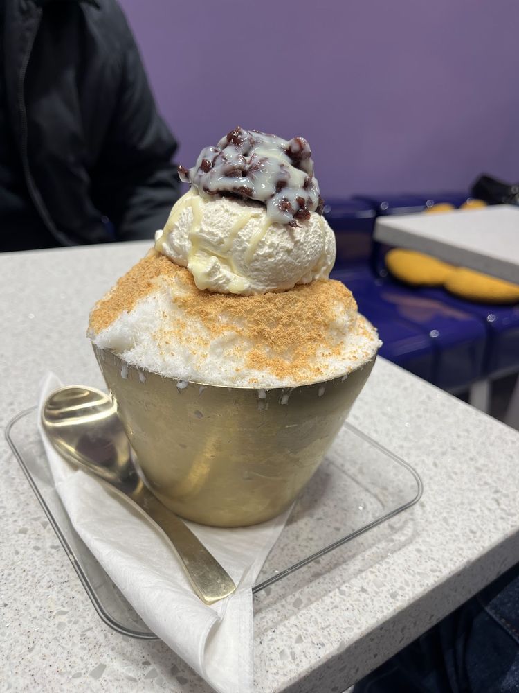 A gold cup of shaved-ice dessert piled with cream and red bean, with a spoon resting on the stone table.
